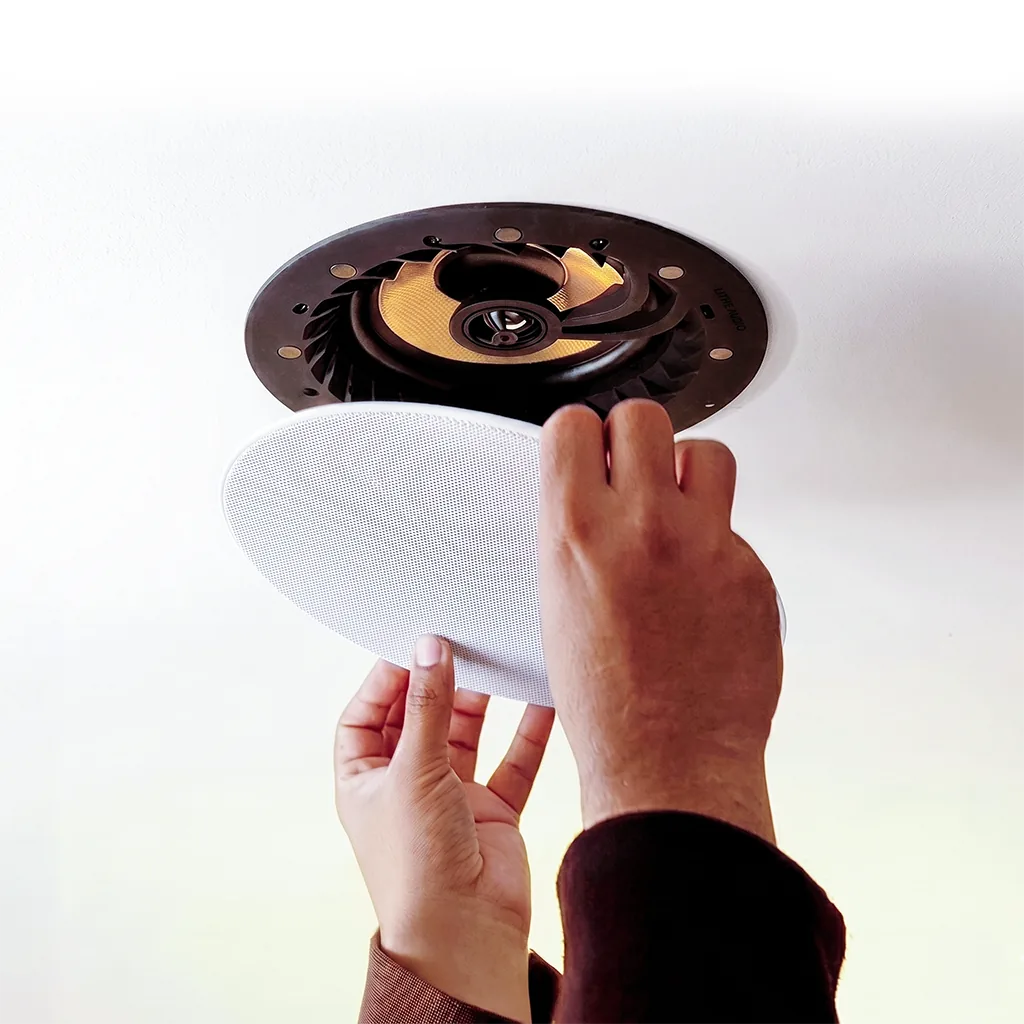 Two hands clipping the magnetic grille onto a Lithe Audio ceiling speaker fitted into a ceiling